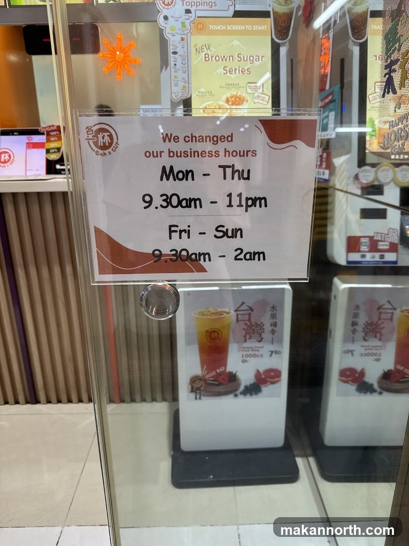 A sign on a glass door displays updated business hours for Each a Cup. The hours are Monday-Thursday from 9:30 AM to 11 PM, and Friday-Sunday from 9:30 AM to 2 AM. Behind the sign, promotional materials for drinks and toppings are visible.