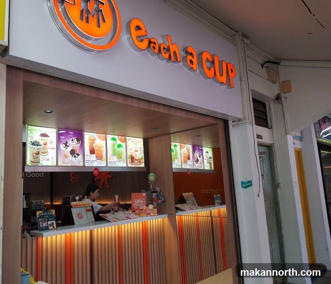 The exterior of a bubble tea shop with the brand name "Each a Cup" displayed prominently above the entrance. The shop features a brightly lit interior with a counter where a staff member is serving customers. Illuminated menu boards showcasing various drinks are displayed above the counter.