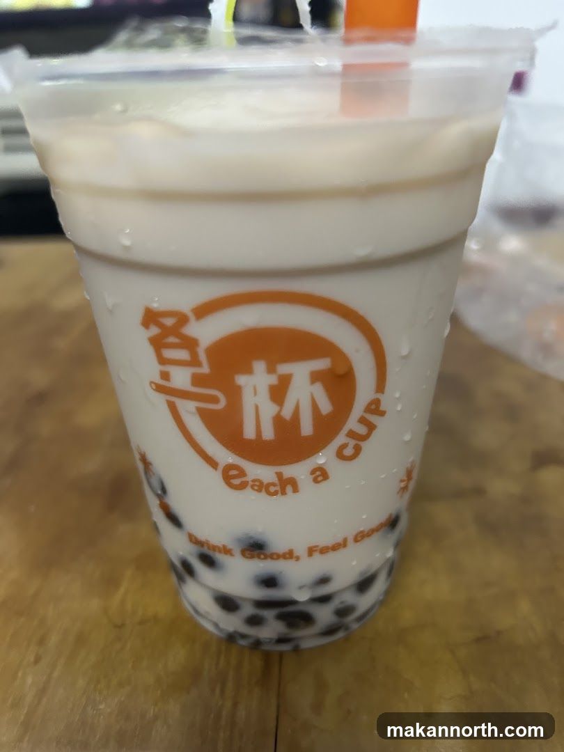 A close-up shot of a bubble tea drink in a clear plastic cup. The cup has a logo with Chinese characters and the text "each a cup" and "Drink Good, Feel Good". The drink is light brown and contains tapioca pearls at the bottom. The cup is covered in condensation.