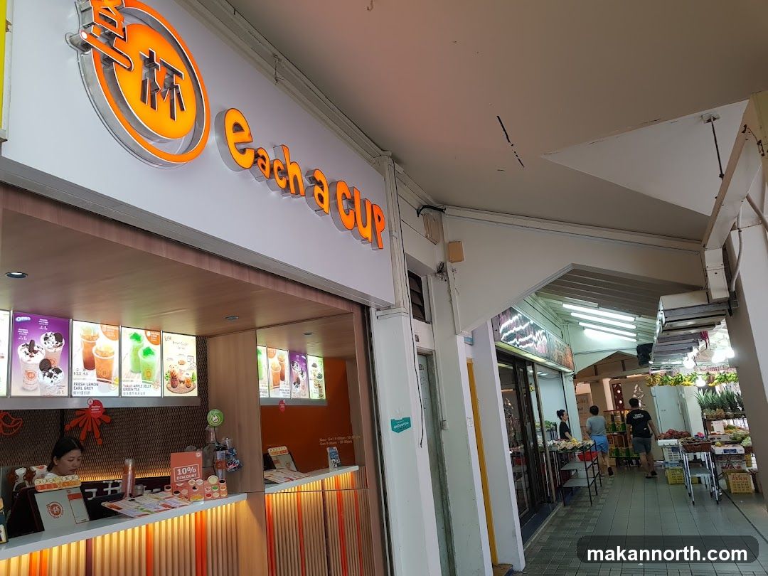 The exterior of a bubble tea shop called Each a Cup. The shop has a prominent orange and white sign with the business name. Inside, illuminated menu boards display various drinks. A staff member is visible behind the counter. The shop is part of a larger walkway with other businesses visible further down.