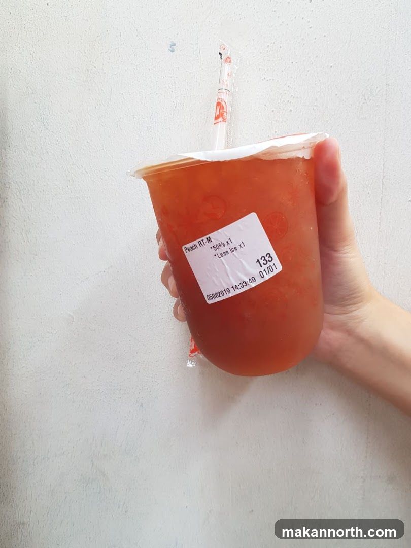 A hand holds a plastic cup filled with a peach-colored beverage. The cup has a white lid and a clear plastic straw with red branding. A white sticker is affixed to the cup, displaying details about the drink.