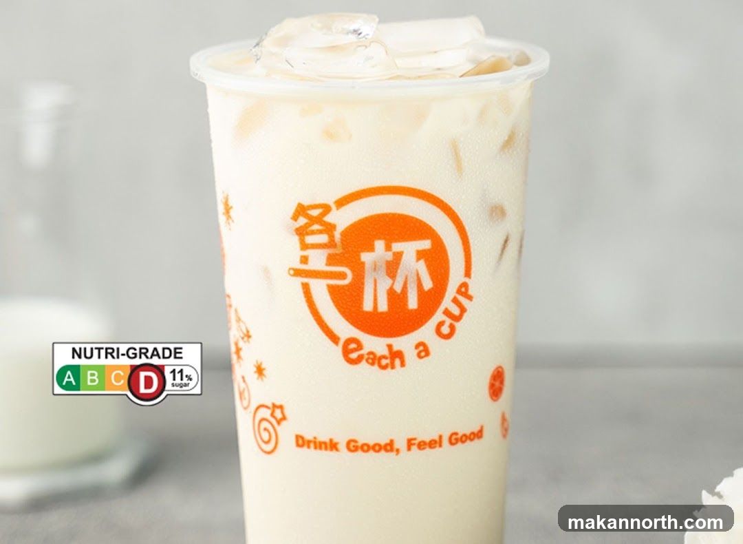 A close-up of a milky beverage in a clear plastic cup with ice. The cup has an orange logo with Chinese characters and the text "Each a CUP" and "Drink Good, Feel Good". A Nutri-Grade label indicating grade D and 11% sugar is visible on the left.