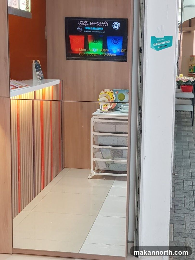 The image shows the interior of a store, with a digital display screen mounted on a wooden wall. The screen shows three colorful cups and text indicating operating hours: Mon-Sat 9:00am-10:30pm and Sun 9:00am-10:00pm. A Deliveroo sticker is visible on the exterior wall. Inside, a counter with orange and wood-paneled accents is reflected in a mirror. A white rolling cart with shelves is also present.