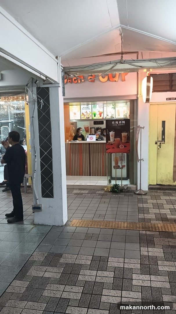 A shop front with a counter and menu displays. Two staff members are visible behind the counter. The shop has orange and white striped paneling. A sign above the counter reads "Bach a Cup". A promotional poster for "Rose Series" drinks is displayed.