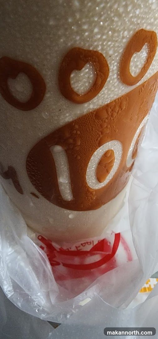 A close-up of a beverage cup with condensation on its surface. The cup has a white textured background with an orange logo featuring circular shapes, some resembling paw prints or hearts. The text "100" is visible in white within the orange design. The cup is partially enclosed in a clear plastic bag, and a red lid or coaster with white text is visible at the bottom.