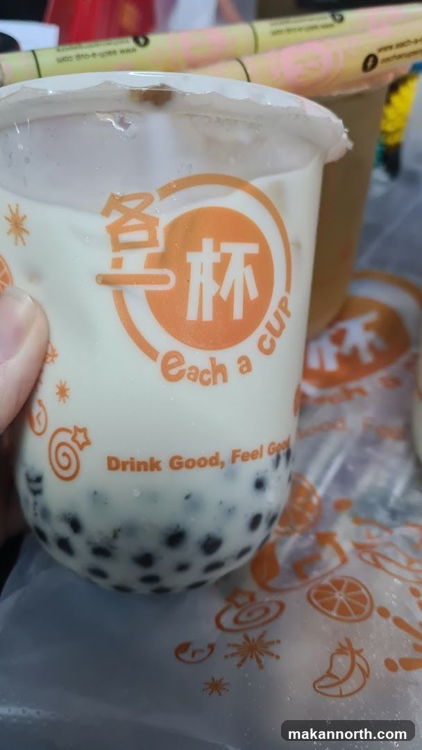 A close-up of a cup of bubble tea with tapioca pearls at the bottom. The cup has an orange logo with Chinese characters and the text "Each a CUP" and "Drink Good, Feel Good". There are other cups and a plastic bag with fruit designs in the background.
