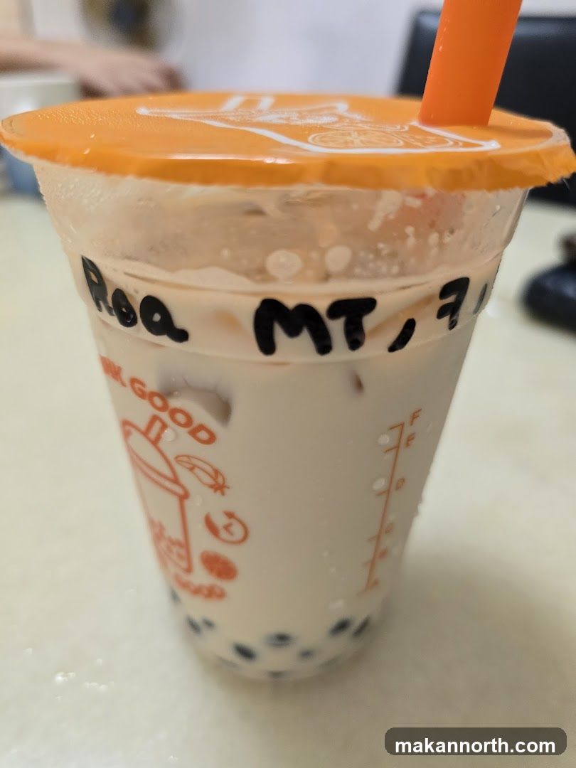 A close-up shot of a bubble tea drink in a clear plastic cup with an orange lid and straw. The cup has condensation on the outside. Black marker writing on the cup reads "Roa MT, 7". The cup has orange branding with icons of a cup, a feather, a clock, and a coffee bean, along with the text "GOOD". There are tapioca pearls at the bottom of the drink.