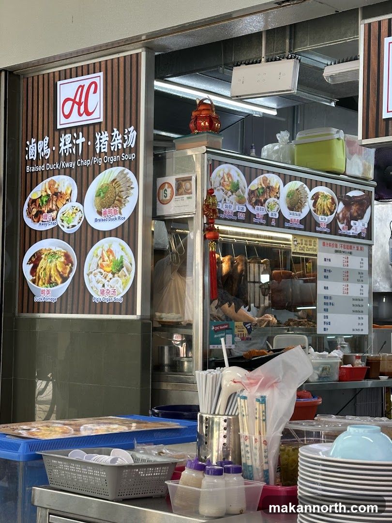A food stall with a sign displaying the business name and various dishes. The sign features images of braised duck, kway chap, duck porridge, and pig's organ soup. Below the images, there is a menu with prices. A person is visible working behind the counter.