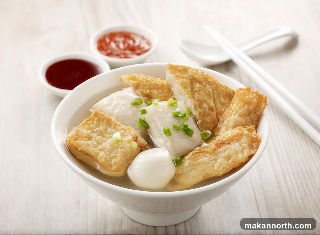 A close-up shot of a bowl of fish ball soup, garnished with chopped green onions. The soup contains various types of fish balls and fried tofu. Two small dipping bowls, one with a red sauce and the other with a dark sauce, are visible in the background, along with a pair of chopsticks and a spoon.
