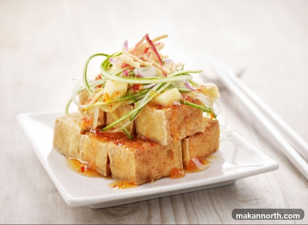 A stack of golden-brown fried tofu cubes sits on a white square plate. The tofu is drizzled with a vibrant orange sauce and topped with a salad of shredded cucumber, red onion, and crispy noodles. White chopsticks rest on the side of the plate.