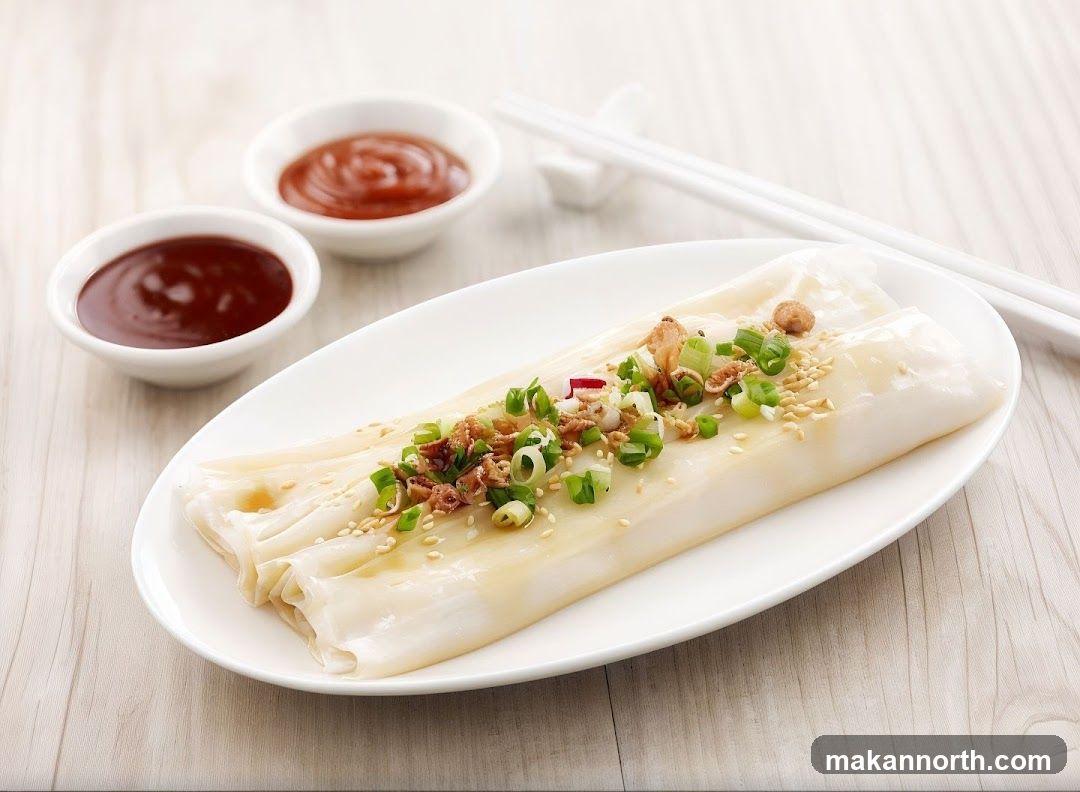 A plate of rice noodle rolls, also known as cheung fun, is served with two small bowls of dipping sauce and chopsticks. The rolls are topped with chopped scallions, sesame seeds, and crispy fried shallots.