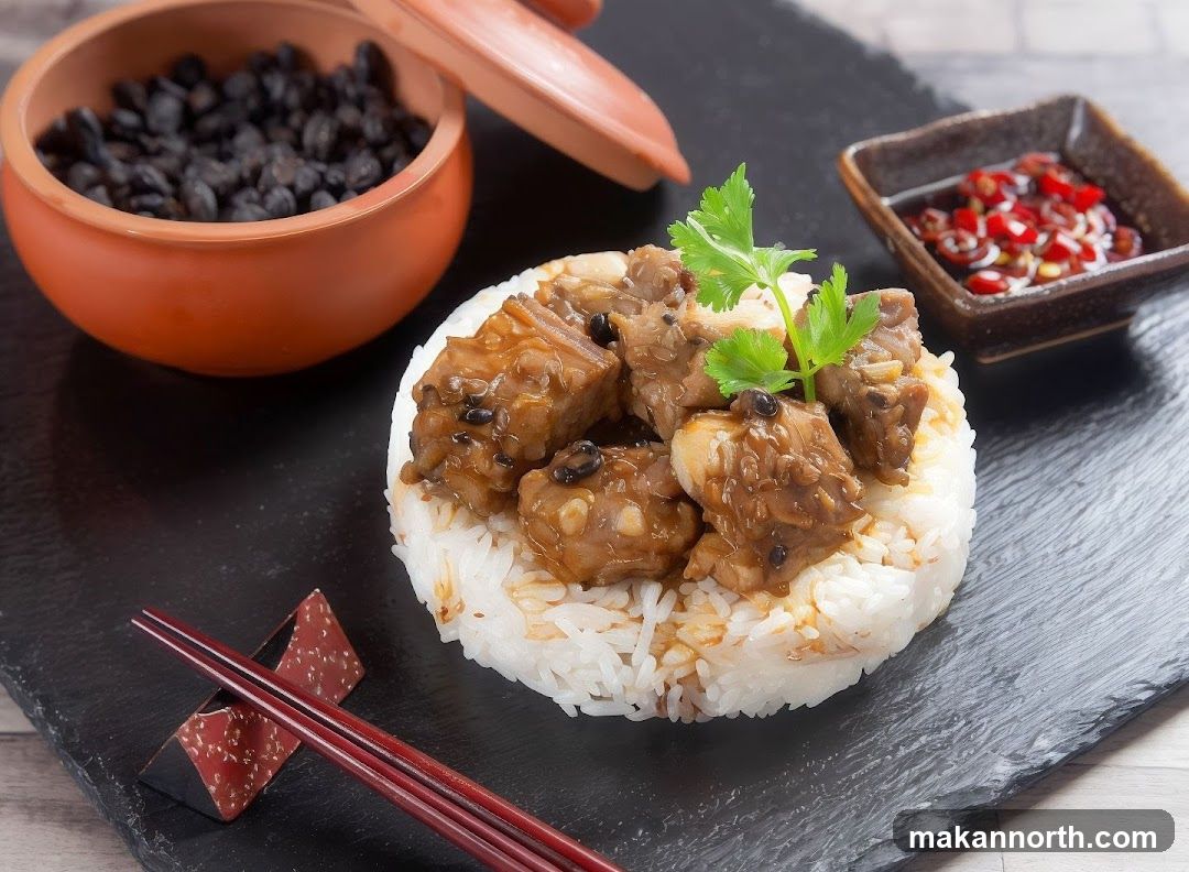 A serving of steamed pork ribs with black beans, garnished with cilantro, is presented on a bed of white rice. A small dish of chili sauce and a bowl of black beans are also visible. Chopsticks rest on a decorative chopstick holder.