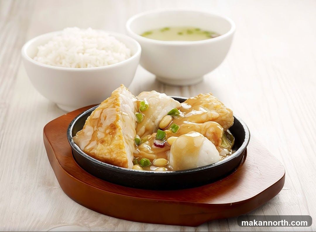 A close-up shot of a sizzling dish served in a black cast-iron pan on a wooden base. The dish contains pieces of white fish, possibly tofu, and other ingredients coated in a glossy brown sauce, garnished with green onions and peanuts. Accompanying the dish are two white bowls, one filled with white rice and the other with a light-colored soup with green specks. The background is a light-colored, textured surface, likely a wooden table.