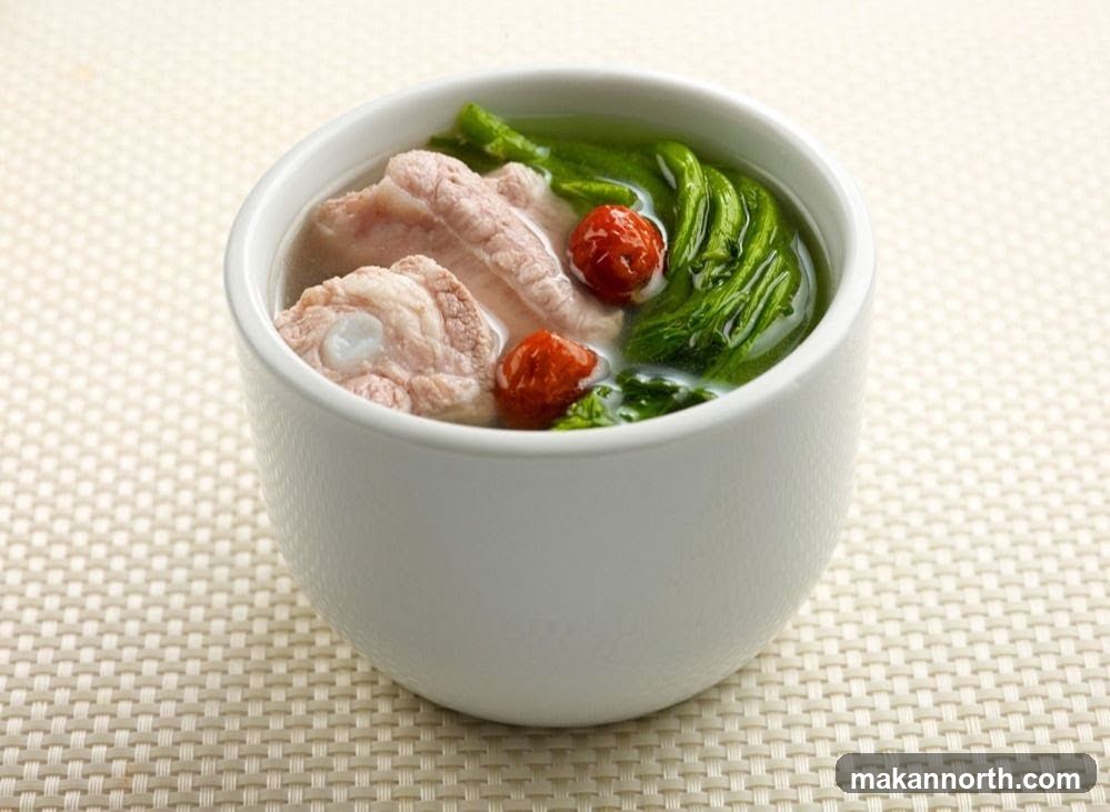 A close-up shot of a white bowl filled with a clear broth soup. The soup contains several pieces of pork ribs, leafy green vegetables, and two red berries. The bowl is placed on a textured, light-colored placemat.