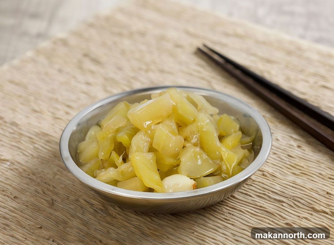 A close-up shot of pickled mustard greens in a metal bowl, served with a pair of dark wooden chopsticks. The greens are chopped into bite-sized pieces and appear to be glistening in a savory sauce. The bowl is placed on a textured placemat, with a blurred background suggesting a simple dining setting.