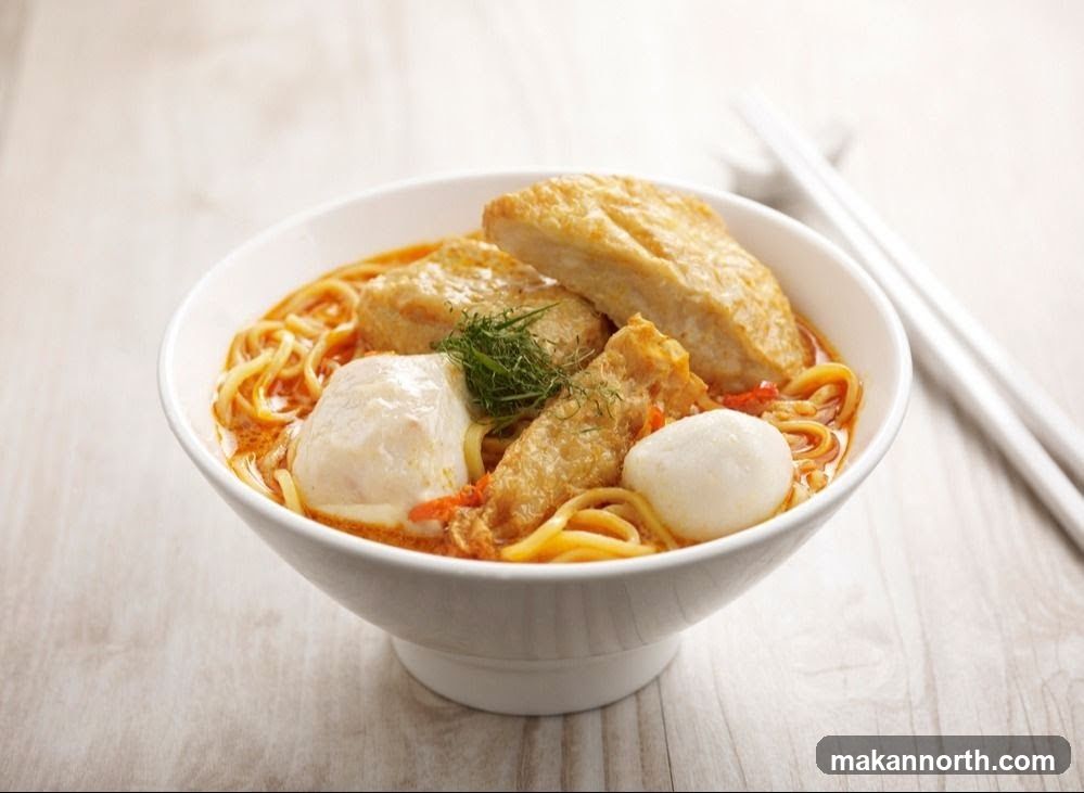 A white bowl filled with noodle soup. The soup has a creamy orange broth and is topped with noodles, fried tofu, fish balls, and a sprig of dill. A pair of white chopsticks rests on the side.