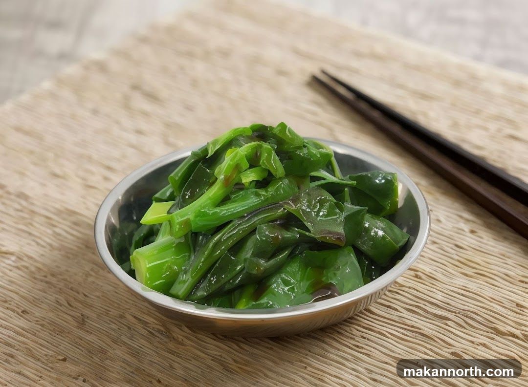 A close-up shot of a bowl of steamed green vegetables, likely Chinese broccoli or similar, drizzled with a dark sauce. The vegetables are glossy and appear tender. A pair of dark wooden chopsticks rests on a woven placemat in the background.