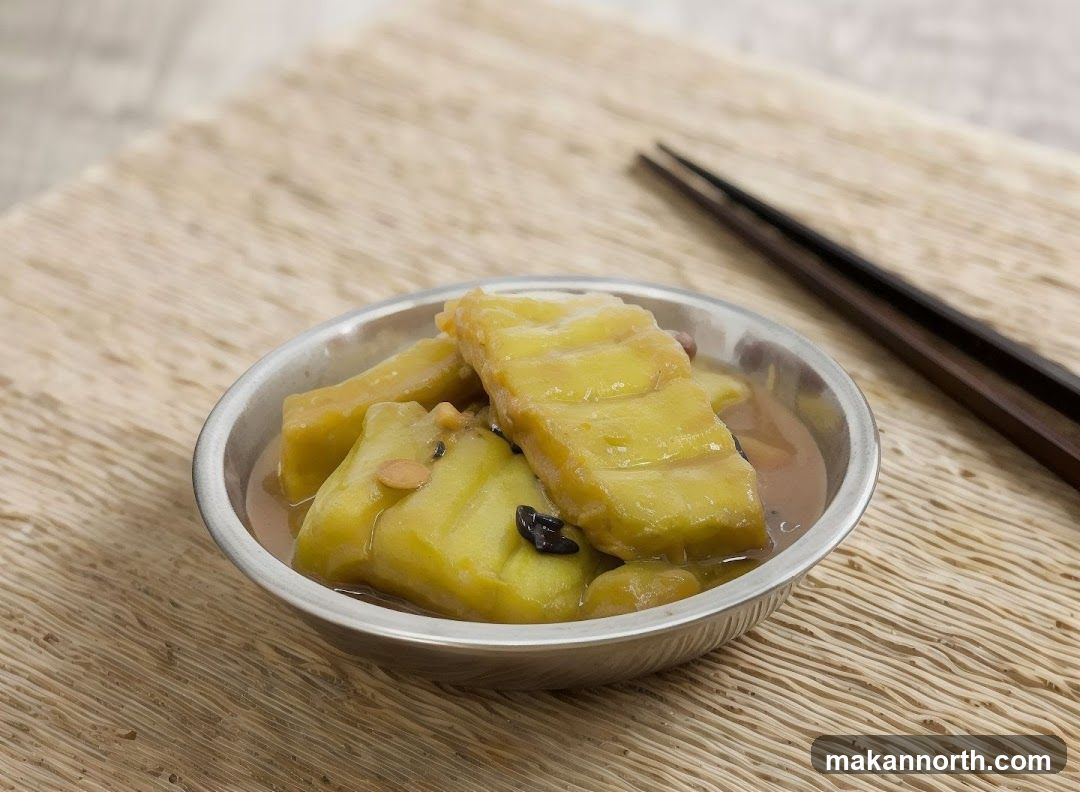 A close-up shot of stewed sweet potatoes in a metal bowl, served with chopsticks on the side. The sweet potatoes are cut into large chunks and appear to be tender and glazed in a light brown sauce. Small dark seeds or spices are visible amongst the sweet potato pieces. The dish is presented on a woven placemat.