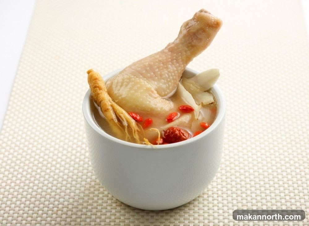A close-up shot of a white bowl filled with chicken soup. A chicken leg is prominently displayed, emerging from the broth. Visible ingredients include a piece of ginseng root, goji berries, and what appears to be slices of ginger or other root vegetables. The soup is served on a textured, light-colored placemat.