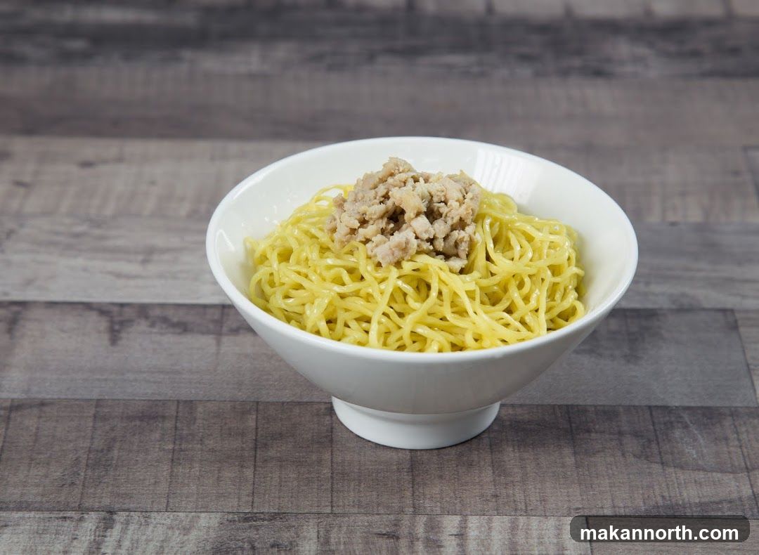 A white bowl filled with yellow noodles and topped with a pile of minced meat. The bowl is placed on a wooden surface.
