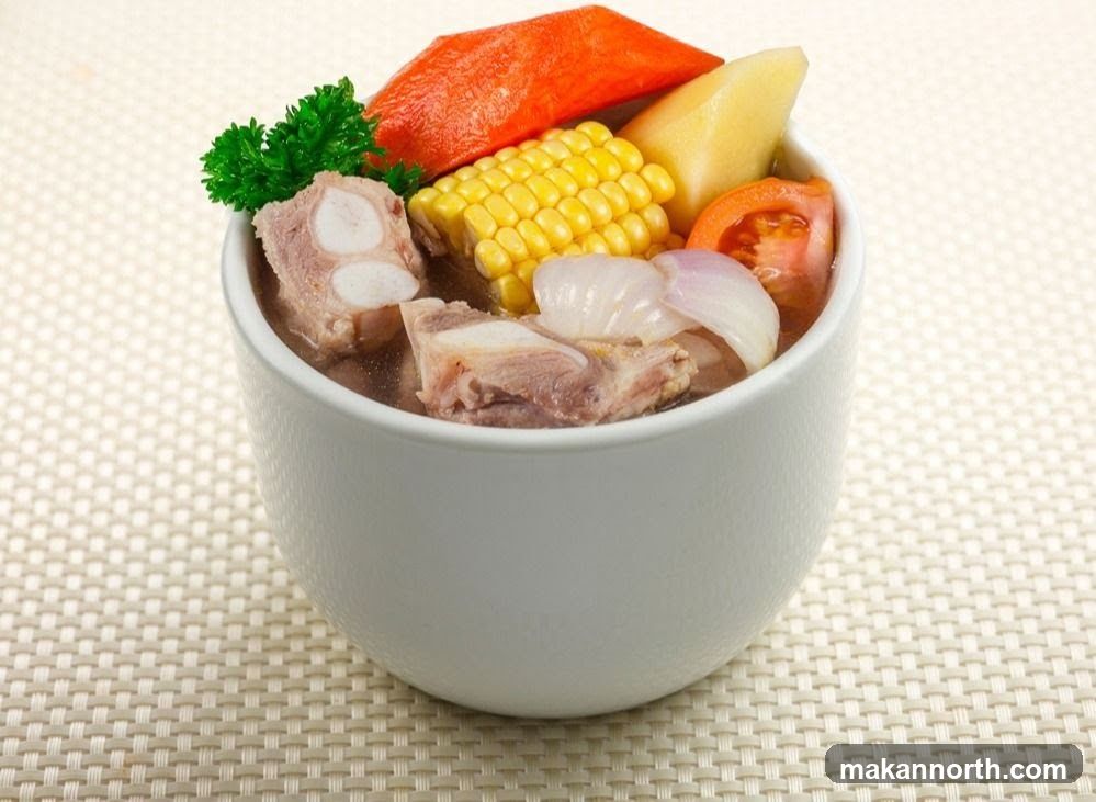 A white bowl filled with a hearty soup containing pork ribs, corn on the cob, sliced tomatoes, onions, a piece of carrot, and a wedge of potato. A sprig of parsley garnishes the top. The bowl sits on a textured placemat.