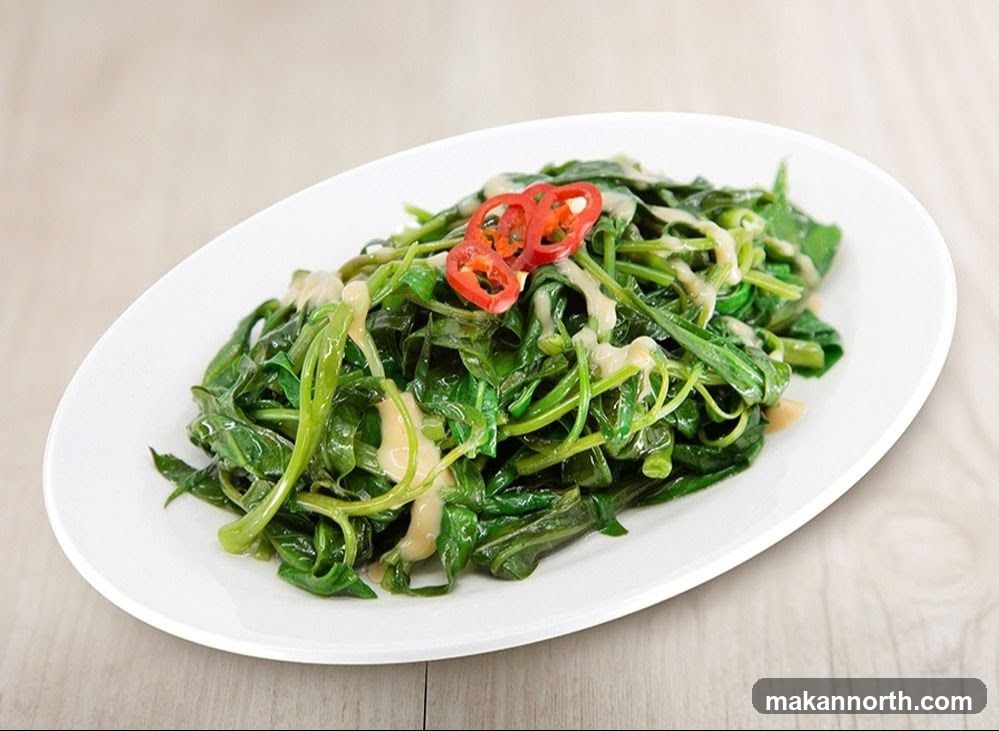 A white oval plate filled with stir-fried water spinach, drizzled with a creamy sauce and garnished with sliced red chili peppers. The dish is presented on a light-colored wooden surface.