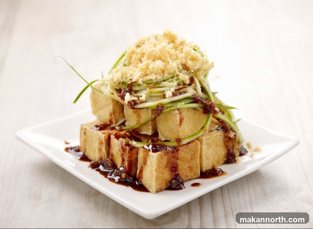 A close-up shot of fried tofu cubes stacked on a white square plate. The tofu is drizzled with a dark sauce and topped with shredded cucumber and a sprinkle of crispy crumbs.