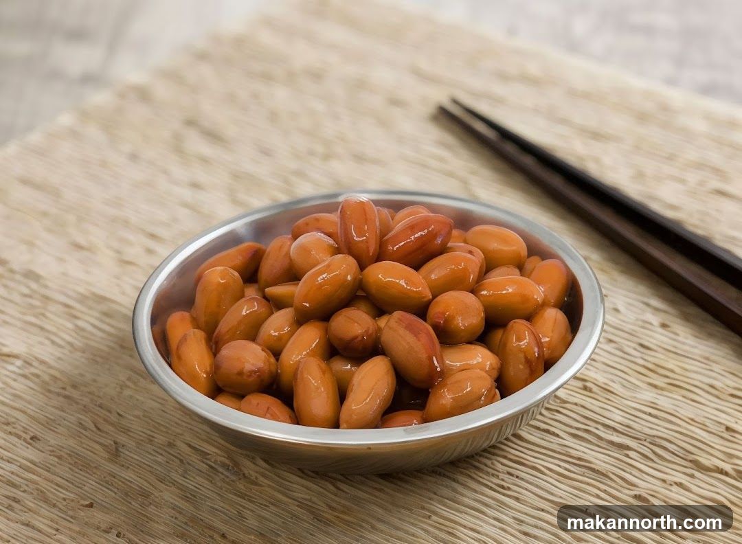 A close-up shot of a metal bowl filled with cooked peanuts. The peanuts are a reddish-brown color and appear to be coated in a light glaze. A pair of dark wooden chopsticks rests to the right of the bowl. The bowl and chopsticks are placed on a textured, light-brown placemat.