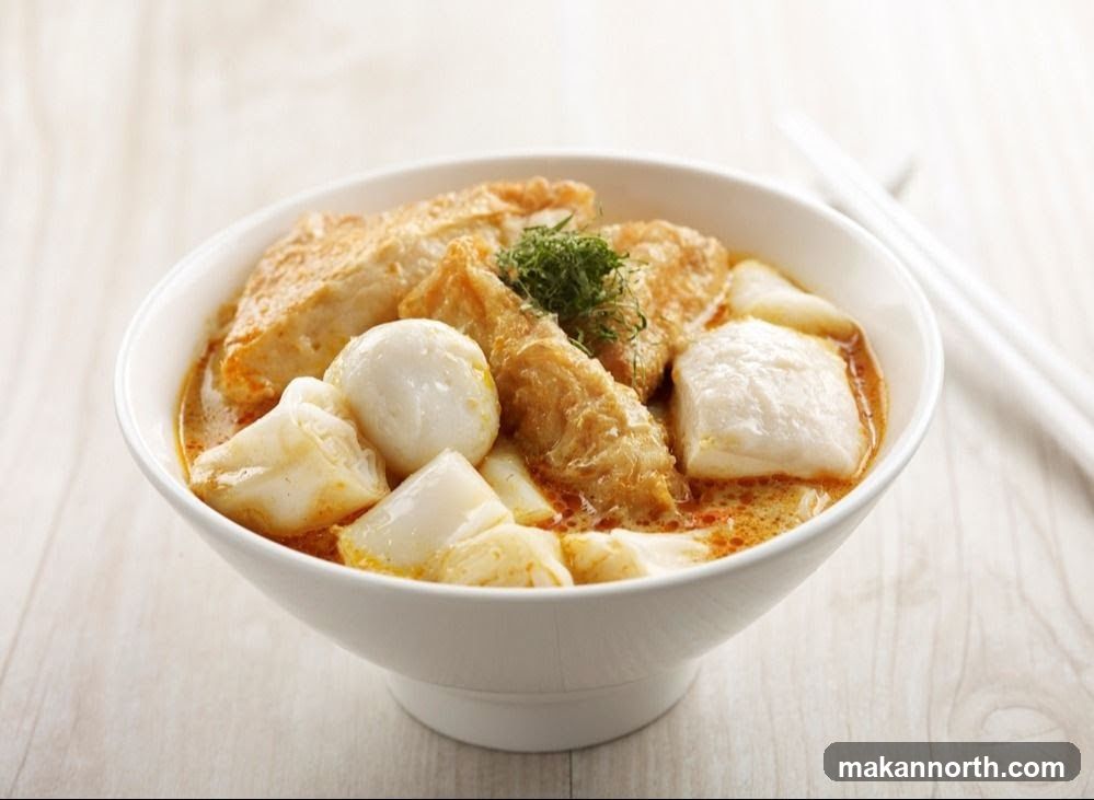 A close-up shot of a bowl of Curry Laksa, a popular Southeast Asian noodle soup. The dish features a rich, orange-hued curry broth filled with various ingredients. Visible components include fried tofu puffs, fish balls, and thick rice noodles. A small garnish of green herbs sits atop the dish. White chopsticks rest on the right side of the bowl, against a light wooden background.