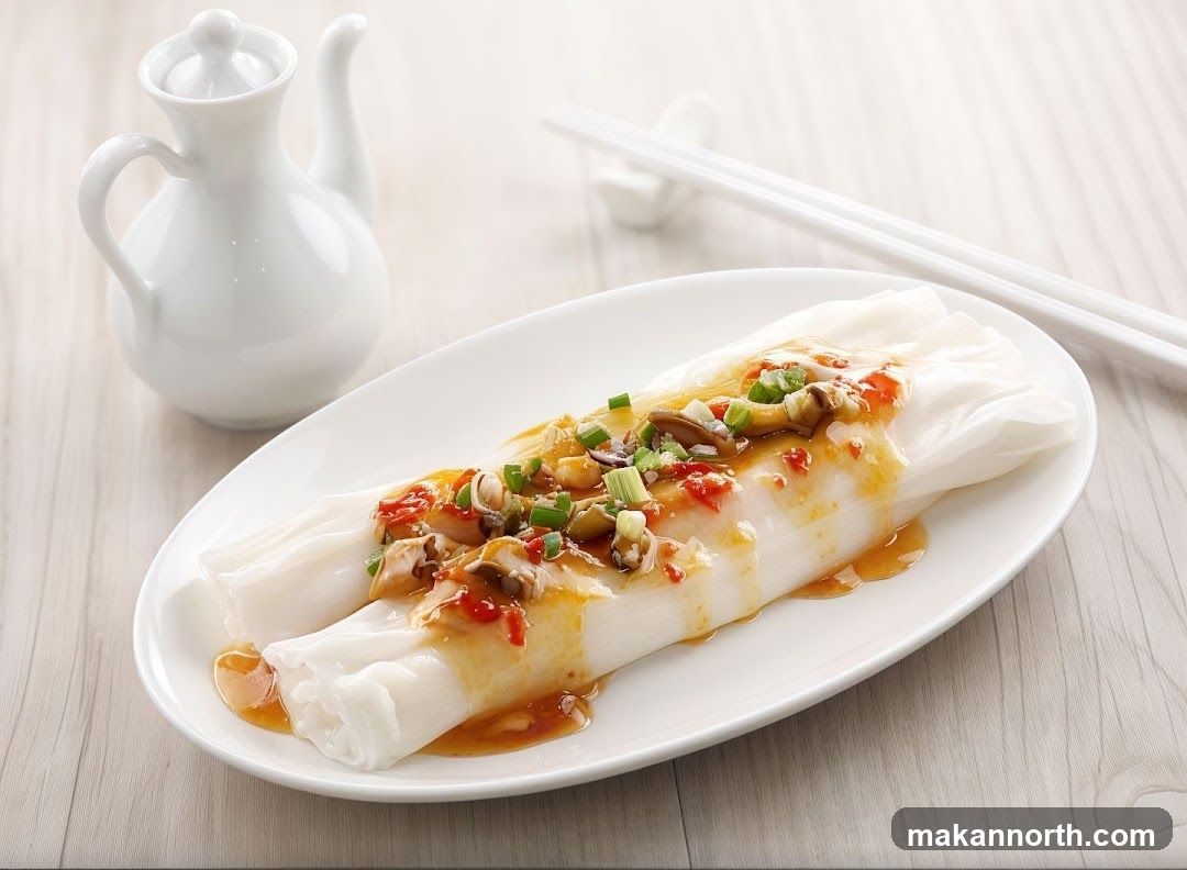 A close-up shot of two steamed rice rolls served on a white oval plate. The rolls are topped with a savory sauce, chopped scallions, chili flakes, and what appear to be small pieces of mushrooms or seafood. A white ceramic sauce pitcher and a pair of white chopsticks rest in the background on a light wooden surface.