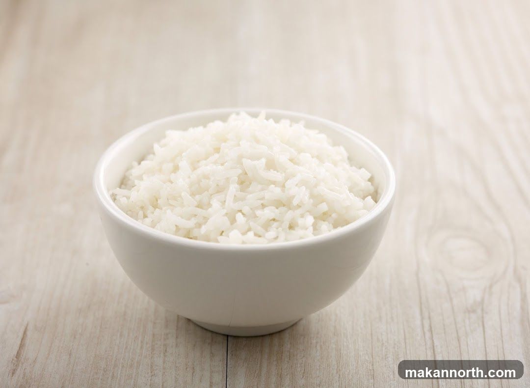 A white bowl filled with fluffy white rice sits on a light-colored wooden surface. The rice is piled high in the bowl, with individual grains visible. The background is softly blurred, with the wood grain texture subtly apparent.