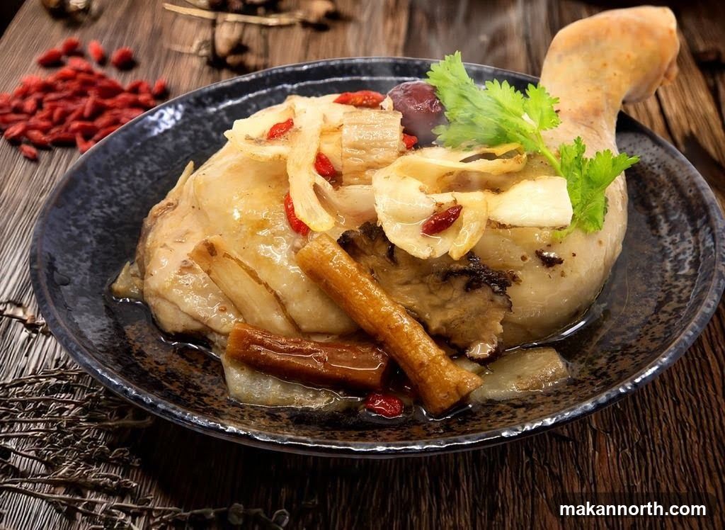 A close-up shot of a whole chicken leg, braised with various herbs and spices, served in a dark ceramic bowl on a wooden surface. The chicken is topped with sliced white root vegetables, red goji berries, and a sprig of cilantro. A pile of goji berries is visible in the background.
