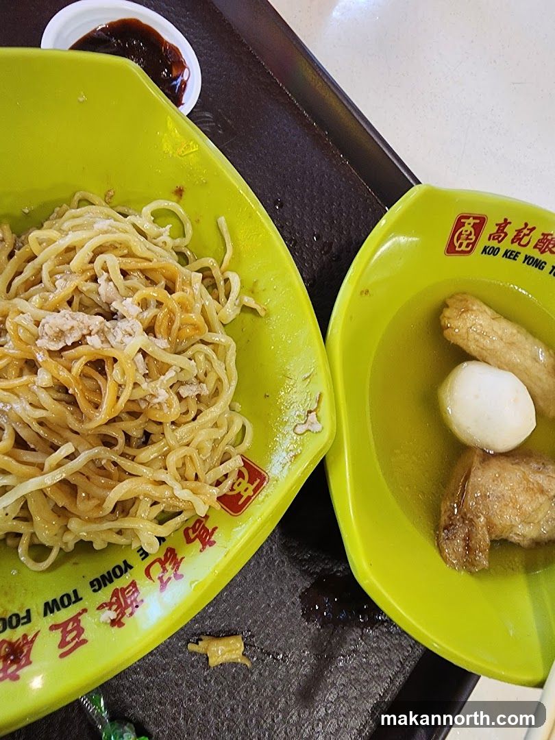 A close-up shot of two lime green bowls on a dark tray. The bowl on the left contains noodles with minced meat. The bowl on the right contains fish balls and other fried items in broth. A small dish of dark sauce is visible in the upper left corner. The bowls have red and yellow branding with Chinese characters and the text "KOO KEE YONG T" and "YONG TOW FOO".