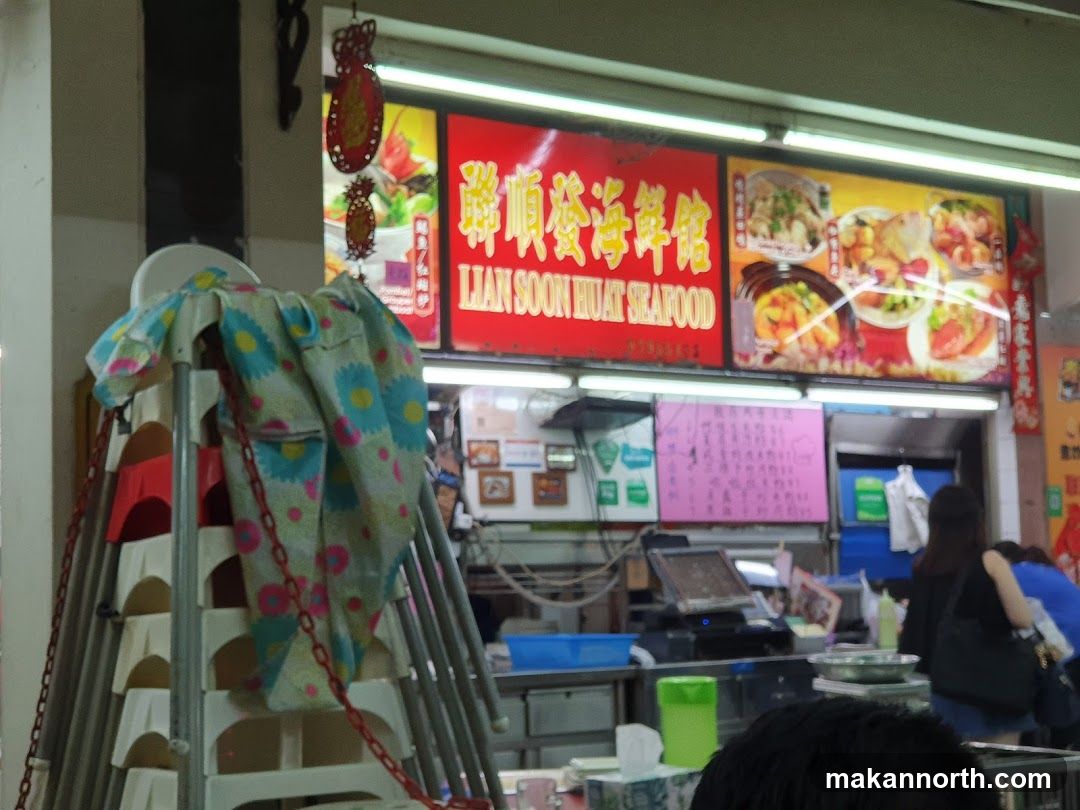 A brightly lit restaurant facade with a large red sign displaying "LIAN SOON SEAFOOD" in English and Chinese characters. The sign is accompanied by images of various seafood dishes. Below the main sign, there are smaller signs and a counter area with a cash register and food preparation equipment. Stacks of chairs are visible to the left of the entrance.