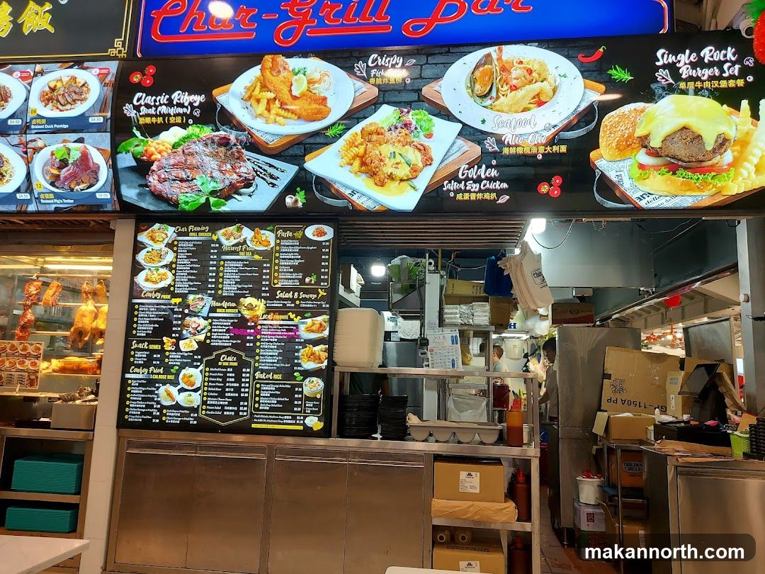 A food stall with a large illuminated sign and menu boards. The sign displays images of various dishes including steak, fish and chips, seafood pasta, and a burger. Below the sign are two large menu boards listing different food categories and items. The stall appears to be busy with staff working in the background.
