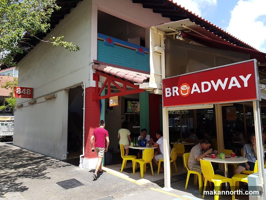 A street view of the Broadway restaurant with a large red sign displaying the name. The restaurant has outdoor seating with yellow chairs and tables. People are seen dining at the tables. The building has a light-colored facade with some colorful accents. A smaller sign with the number 848 is visible on the left side of the building.