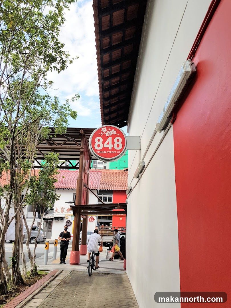 A red and white building with a sign that says "848 YISHUN STREET 81" and a cherry blossom design. The building has a tiled roof and a white light fixture on the exterior wall. In the background, there are other buildings and people walking and cycling.