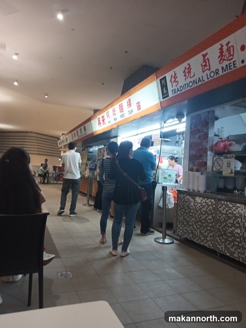 A food stall with a sign that reads "TRADITIONAL LOR MEE" and Chinese characters. People are queuing to order. The stall appears to be in a food court or hawker center.