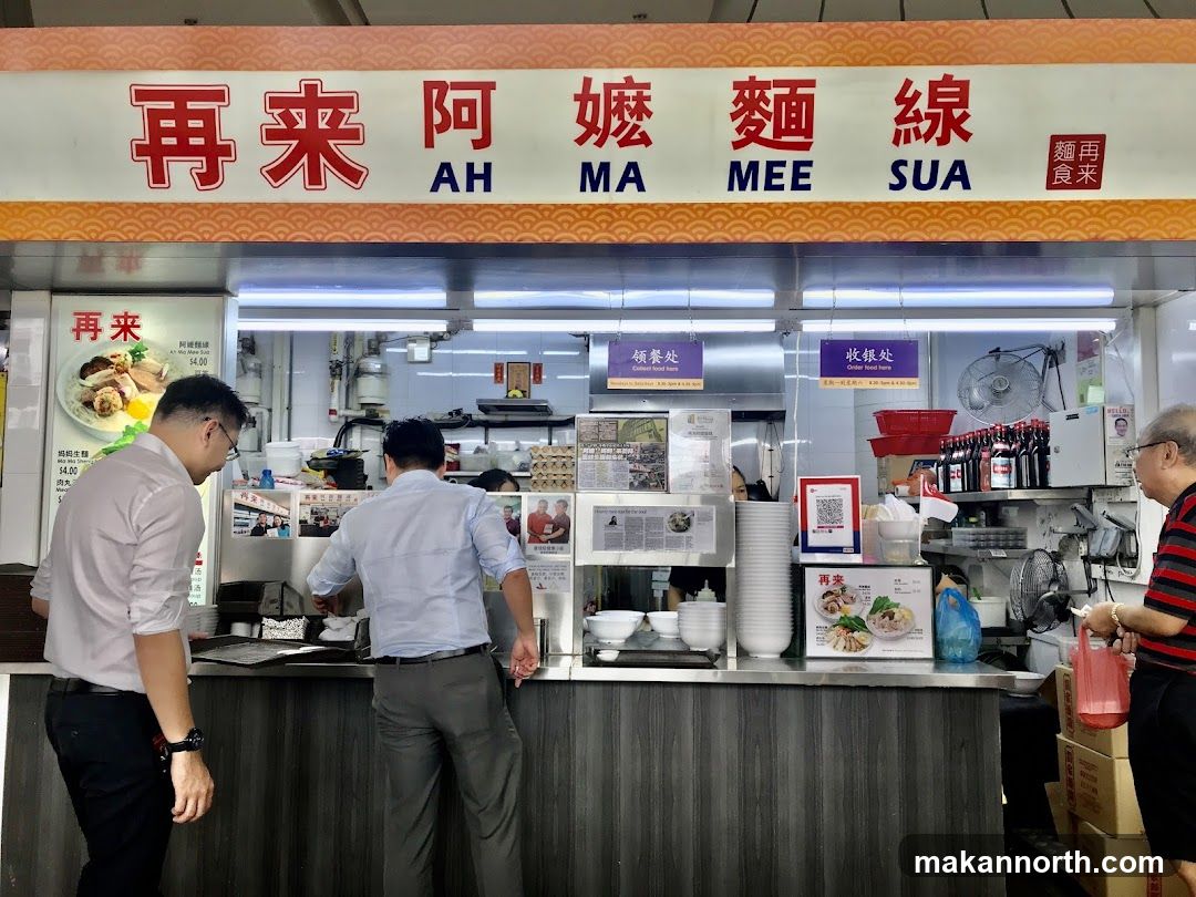 A food stall with a prominent sign reading "再来阿嬤麵線 AH MA MEE SUA". The stall has a counter where customers can order and collect food. Several people are visible, including staff and customers. There are stacks of bowls, bottles of sauces, and food displayed on the counter.