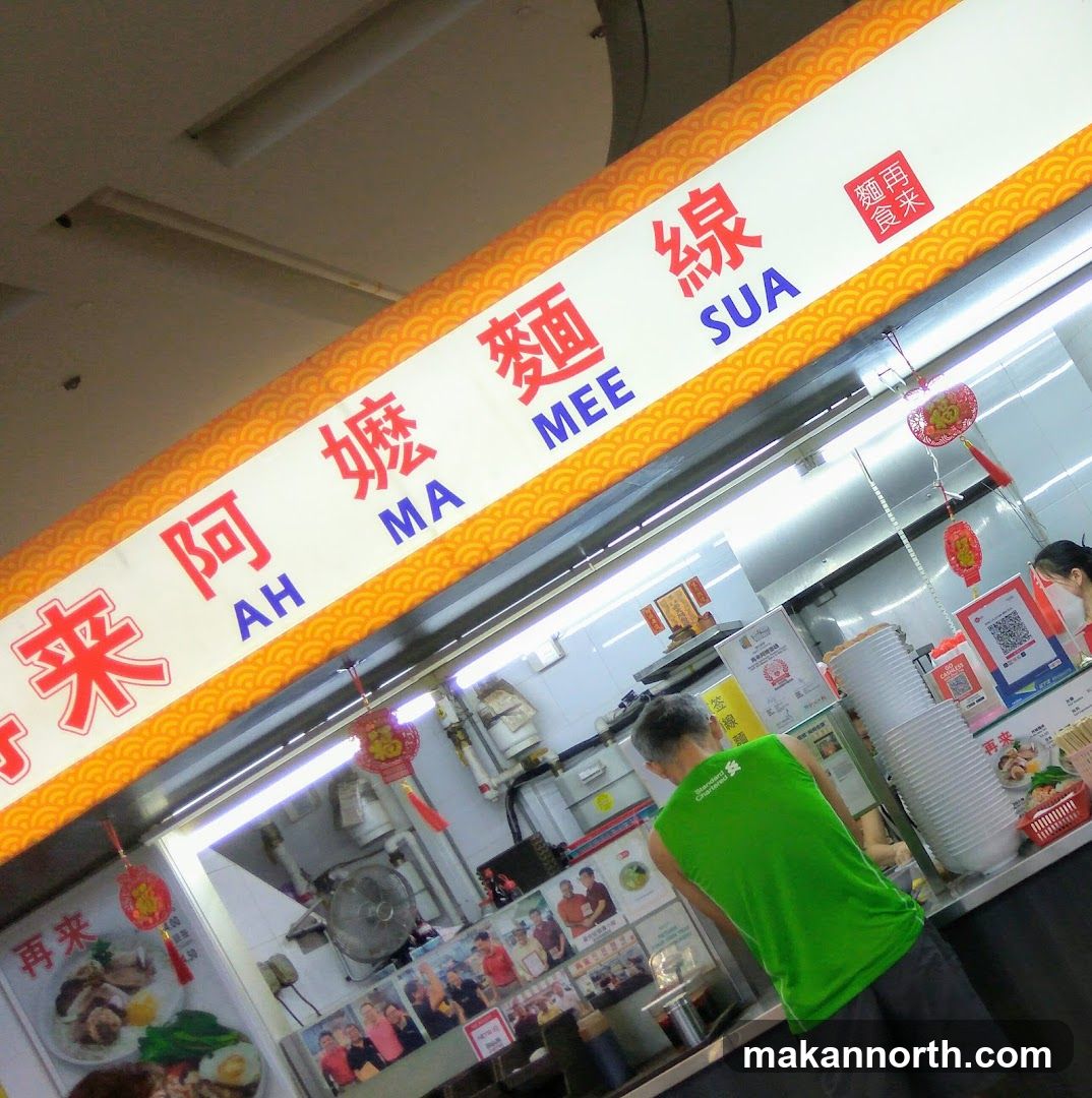 A food stall with a prominent sign displaying "Ah Ma Mee Sua" in Chinese and English. The stall appears to be in a hawker center or food court, with a person in a green shirt working behind the counter. There are various food pictures and decorations visible.