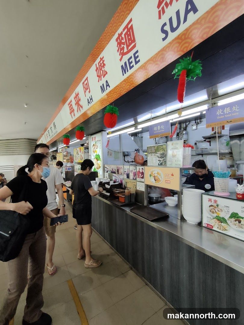 A food stall with a prominent sign displaying "AH MA MEE" in English and Chinese characters. The stall appears to be busy with customers ordering and staff preparing food. Decorative red strawberry-like lanterns hang from the ceiling. Signs indicate "Order food here" and "收银处" (cashier).