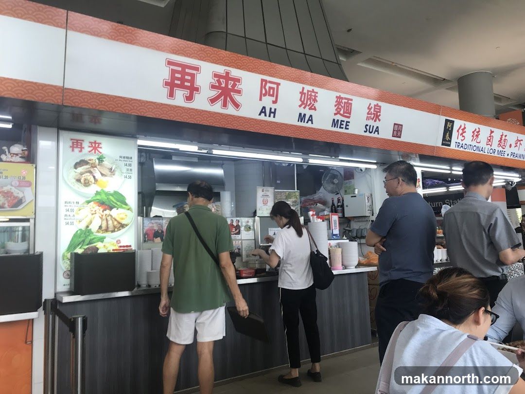 A food stall with a prominent sign displaying "Ah Ma Mee Sua" in both Chinese and English. The stall offers traditional Lor Mee and Prawn dishes. A menu board shows prices for various noodle dishes. Several customers are seen queuing and ordering at the counter.