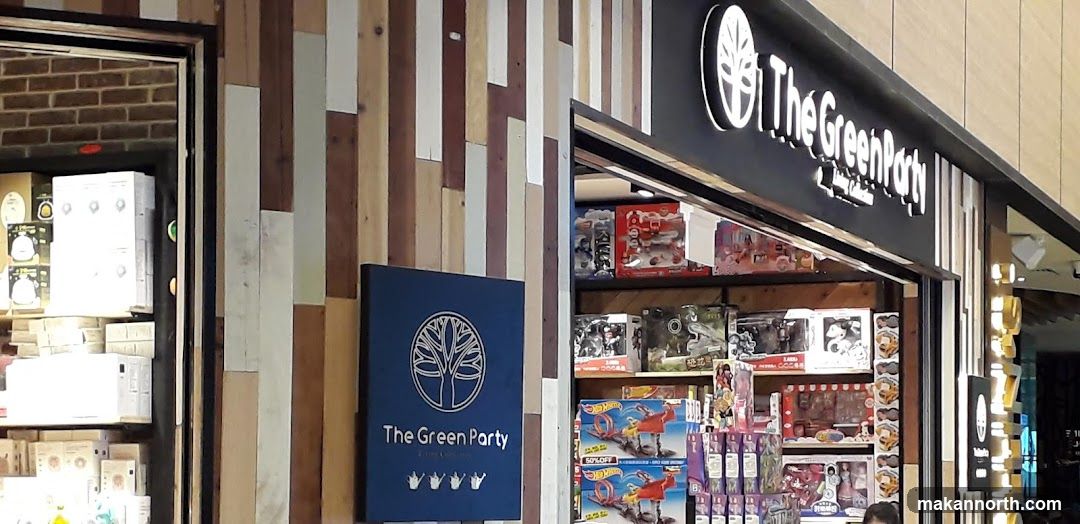 The exterior of a shop called "The Green Party" is visible. The shop front has a dark sign with the store name and a tree logo. The interior of the shop is filled with various toys and merchandise, including action figures, toy cars, and dolls. The walls are decorated with a pattern of vertical wooden planks in different colors.