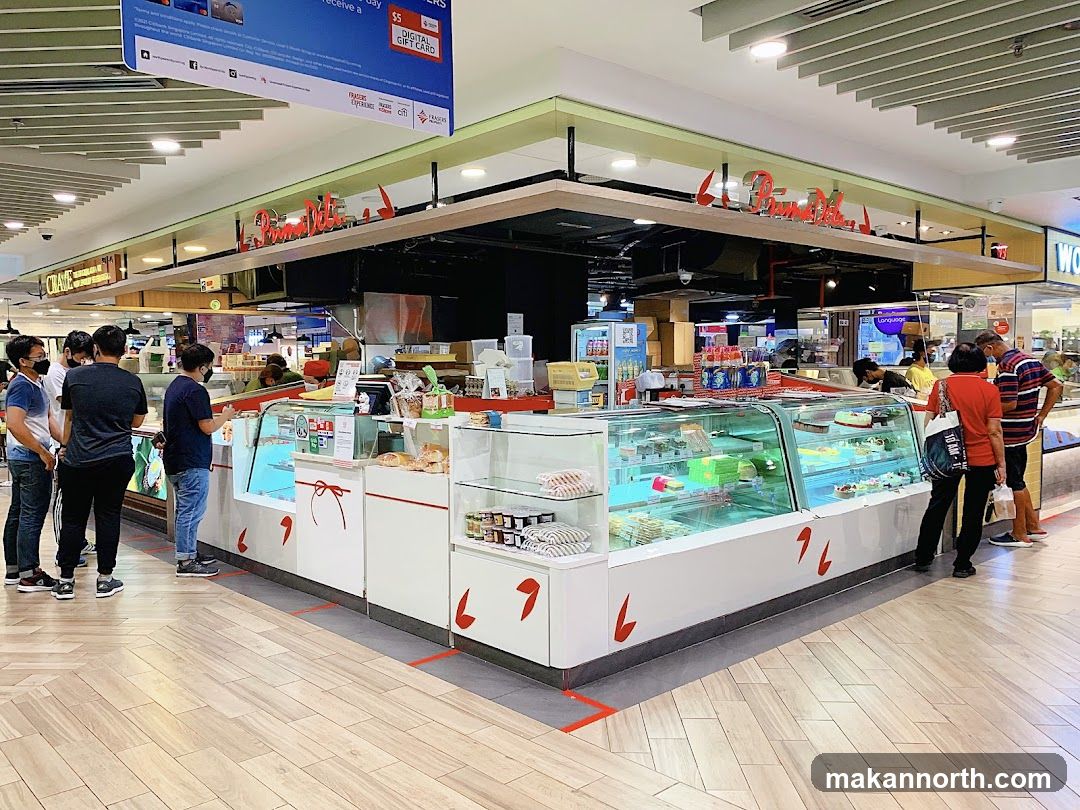 A brightly lit bakery shop front with display cases filled with cakes and pastries. Customers are seen queuing and browsing the selection. The shop's name is visible in red lettering above the counter.