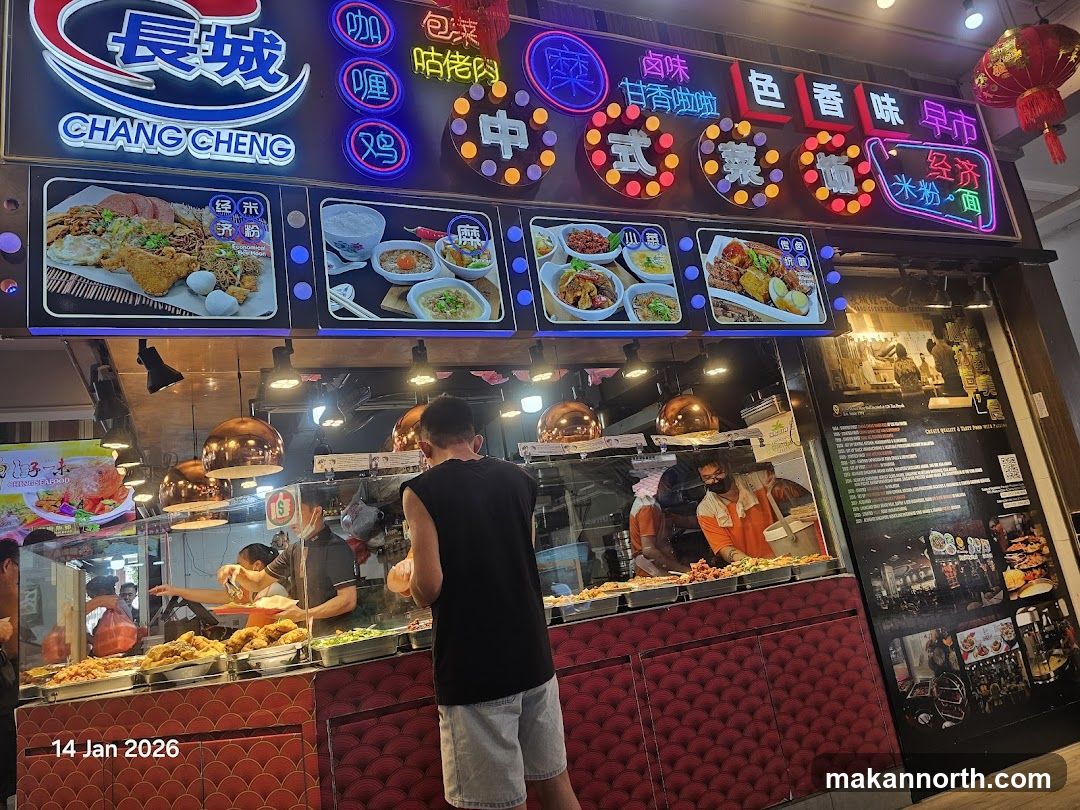A brightly lit restaurant storefront with a large neon sign displaying the name "CHANG CHENG" in English and Chinese characters. Below the sign are illuminated display panels showcasing various dishes. Inside, staff members are visible preparing food behind a counter filled with various food items. The lower part of the storefront features a decorative red patterned facade.