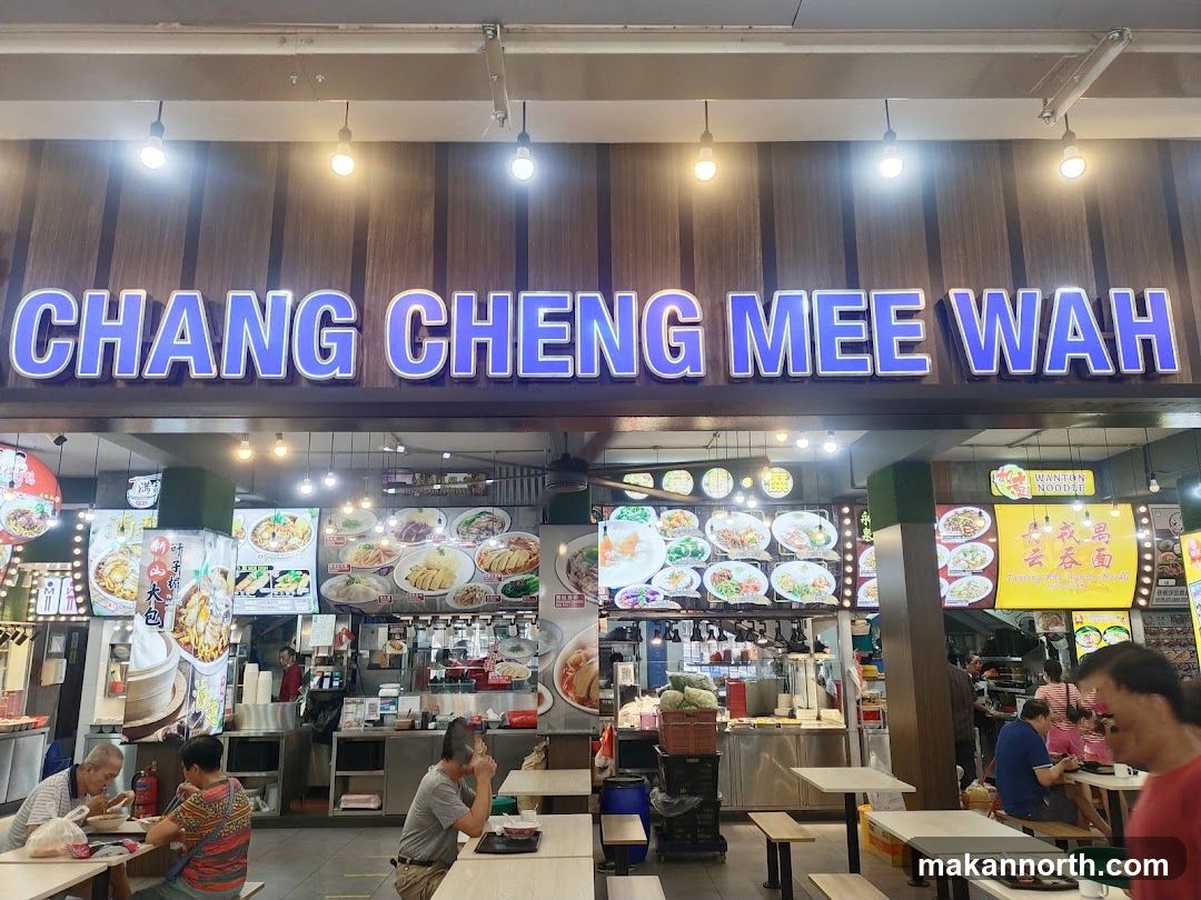 A brightly lit storefront with a large blue neon sign that reads "CHANG CHENG MEE WAH". Below the sign, a food stall displays numerous pictures of various dishes on illuminated boards. People are seated at tables in front of the stall, some eating and others conversing. The interior of the stall shows food preparation areas and counters.