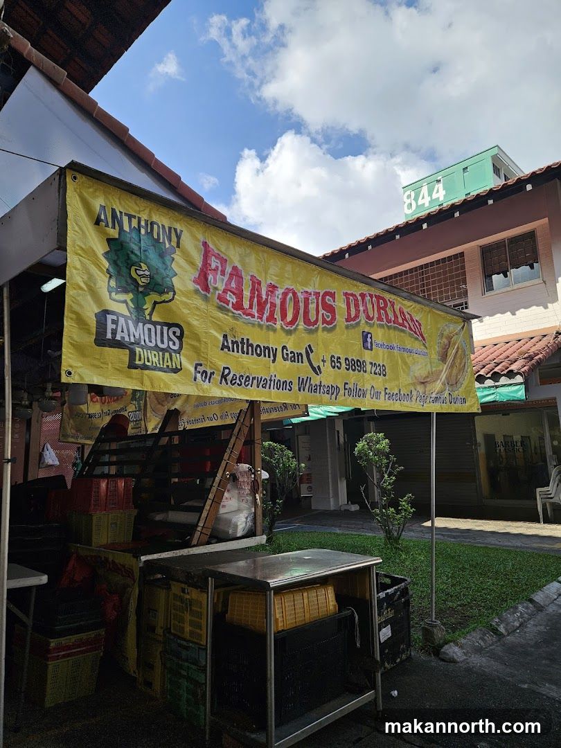 A yellow banner with red and black text reads "ANTHONY FAMOUS DURIAN". Below this, it says "Anthony Gan + 65 9898 7238 For Reservations Whatsapp Follow Our Facebook Page Famous Durian". The banner is hung outside a stall with crates of durians and other produce.