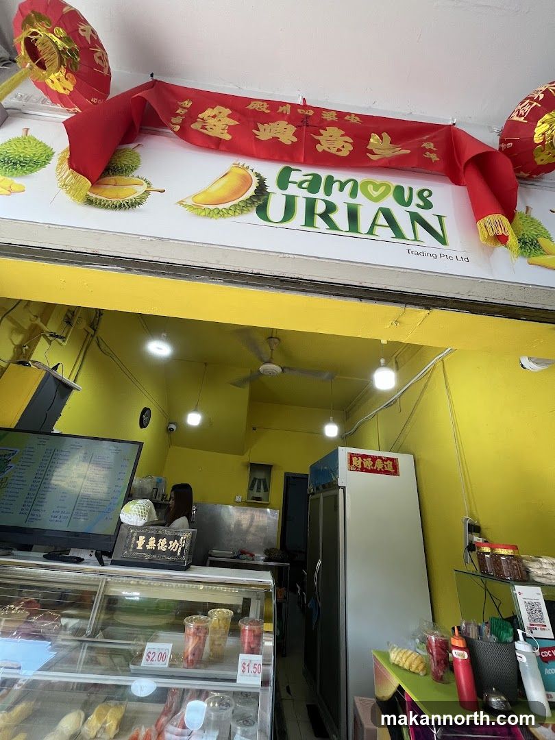 The exterior of a durian shop with a bright yellow facade. A large sign above the entrance reads "Famous Durian" with images of durians. Red lanterns and a red banner with Chinese characters hang above the sign. Inside, a display case shows durian products and other fruits. A menu is visible on a screen to the left.